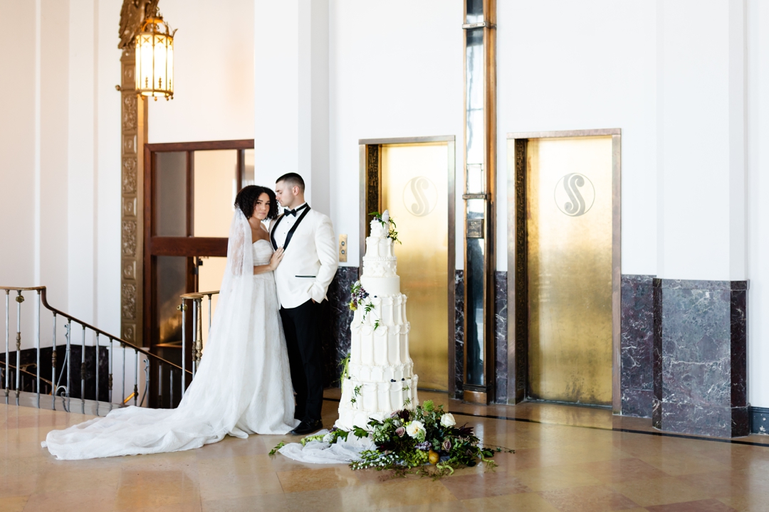 Life-size wedding cake at The Noble – A towering white wedding cake with sculptural piping, fresh greenery, and sugared fruit, standing in front of gold elevator doors.