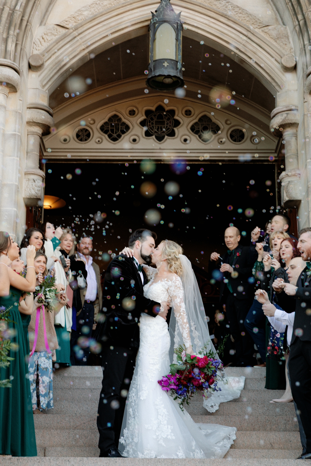 wedding gallery photo of bride and groom kissing on church steps while guests cheer and bubbles floating in the air