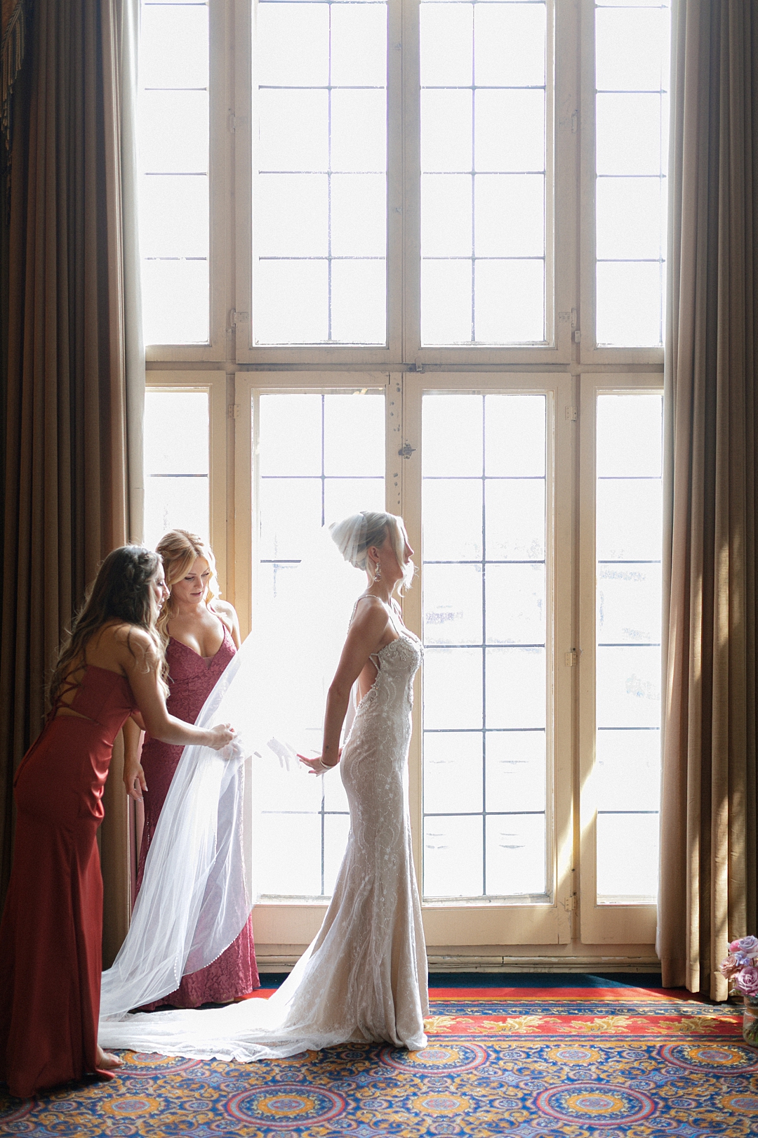 Bride standing in soft window light while her veil is adjusted, creating a cinematic wedding moment