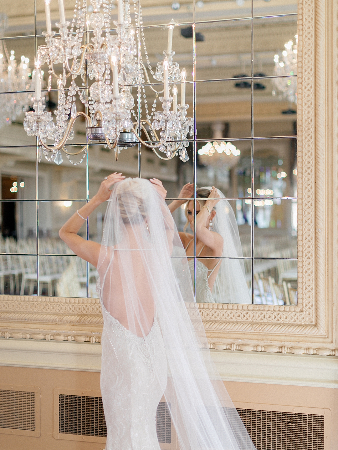Bride adjusting her veil in front of a mirrored wall with chandelier reflections